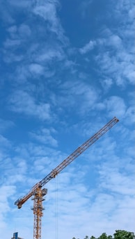 A large construction crane is prominently featured against a bright blue sky with scattered, wispy clouds. The crane is tall and yellow, stretching across the sky. Below the crane, there is a hint of buildings and treetops.