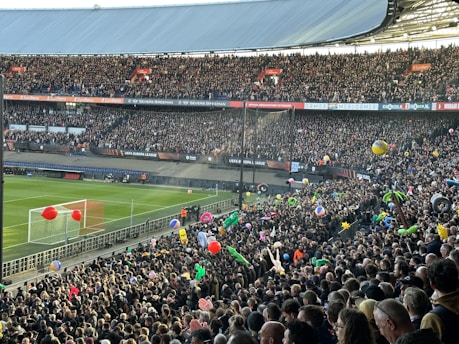 A crowded football stadium filled with enthusiastic fans. Many are holding colorful inflatable beach balls and other objects. The vibrant scene is set against a clear view of the pitch and goalposts. Banners and signs for the UEFA Europa League are prominently displayed, creating a lively and festive atmosphere.