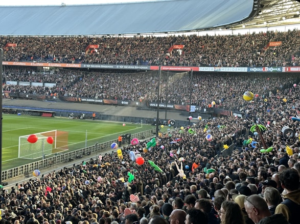 A crowded football stadium filled with enthusiastic fans. Many are holding colorful inflatable beach balls and other objects. The vibrant scene is set against a clear view of the pitch and goalposts. Banners and signs for the UEFA Europa League are prominently displayed, creating a lively and festive atmosphere.