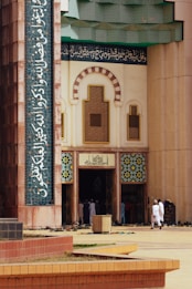 The entrance of a mosque with intricate geometric and floral patterns on the tiles. Arabic calligraphy adorns the facade. People in traditional attire gather at the entrance, with numerous shoes left on the ground outside.