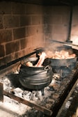 A rustic cooking scene with a black cast iron pot placed on a hearth. The pot, filled with bones and vegetables, is simmering amidst grey ashes, and another pot can be seen in the background. The surrounding bricks are darkened with soot, evoking an old-world kitchen atmosphere.