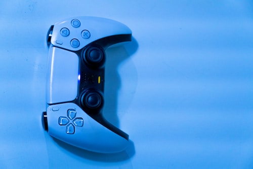 A gaming controller is placed against a blue background. The controller features dual analog sticks, a touchpad, and various buttons including directional arrows and shape symbols. The lighting casts a cool blue hue over the scene, accentuating the sleek design of the controller.