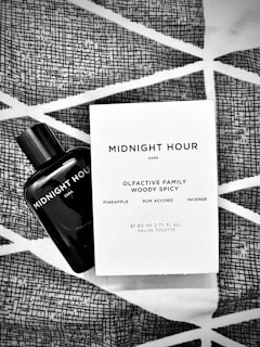 A black bottle of Zara's 'Midnight Hour' fragrance is placed alongside its packaging on a fabric with geometric patterns. The packaging details the olfactive family as woody spicy, with notes of pineapple, rum accord, and incense.