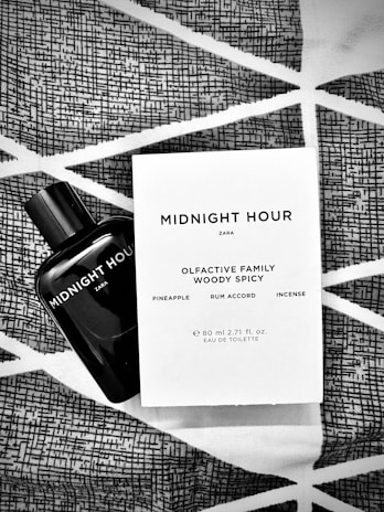 A black bottle of Zara's 'Midnight Hour' fragrance is placed alongside its packaging on a fabric with geometric patterns. The packaging details the olfactive family as woody spicy, with notes of pineapple, rum accord, and incense.