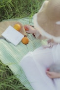 A serene picnic scene with a light green checkered blanket laid on grass, featuring a straw hat and a woven bag. Oranges are placed alongside a white book, and the hand of a person is reaching towards one of the fruits.