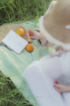 A serene picnic scene with a light green checkered blanket laid on grass, featuring a straw hat and a woven bag. Oranges are placed alongside a white book, and the hand of a person is reaching towards one of the fruits.