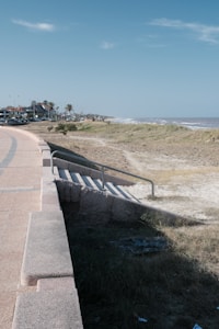 A coastal scene featuring a promenade with a stone pathway and railing running parallel to the beach. The shoreline is lined with grass and dunes, leading to a view of the ocean with gentle waves. In the background, there are residential houses and some palm trees lining the road.