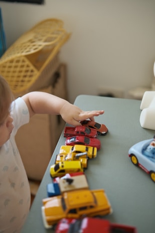 A child reaches towards a lineup of colorful toy cars on a table, with each car varying in size and color. In the background, a wicker basket is partially visible, adding a casual, domestic atmosphere.