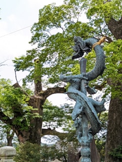 A detailed artistic sculpture of a twisting dragon with intricate scales, wrapped around a vertical pole or staff. The sculpture is placed outdoors, surrounded by lush, green trees and a bright sky. The dragon is depicted with a dynamic posture and a fierce expression.