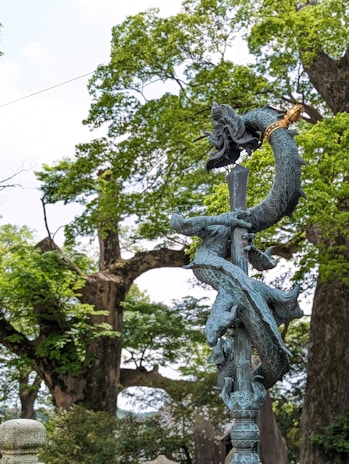 A detailed artistic sculpture of a twisting dragon with intricate scales, wrapped around a vertical pole or staff. The sculpture is placed outdoors, surrounded by lush, green trees and a bright sky. The dragon is depicted with a dynamic posture and a fierce expression.