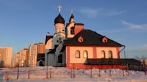 A beautiful Orthodox church with distinctive black domes and crosses on top is bathed in warm sunlight. The building is surrounded by snow and set against a clear blue sky. In the background, modern high-rise buildings can be seen.
