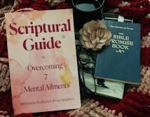 A book titled 'Scriptural Guide to Overcoming 7 Mental Ailments' lies next to a pair of black-rimmed glasses, a large beige flower, and another book titled 'The Bible Promise Book.' These items are placed on a textured, multicolored knitted fabric, and there is a black cup next to the items.
