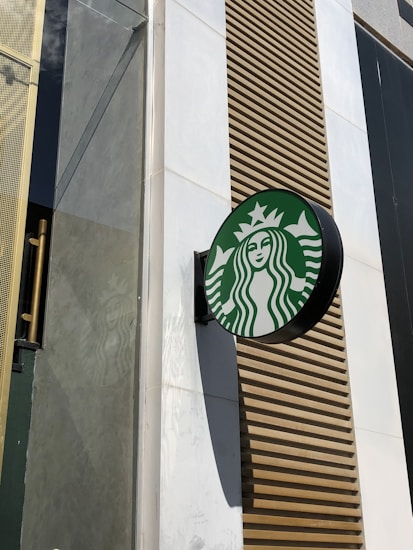 A round sign with a green and white logo featuring a twin-tailed mermaid is mounted on the exterior wall of a building. The building facade includes a mix of materials including glass, metal, and a wood-like texture. The logo casts a shadow on the wall, indicating sunlight.