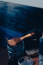 A wooden-handled paintbrush rests on a transparent container that appears to be filled with water. Next to it, there is a well-used sponge and a bottle, suggesting the setting is an artist's workspace. The background features blurry, dark blue shades that enhance the artistic atmosphere.