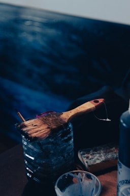 A wooden-handled paintbrush rests on a transparent container that appears to be filled with water. Next to it, there is a well-used sponge and a bottle, suggesting the setting is an artist's workspace. The background features blurry, dark blue shades that enhance the artistic atmosphere.