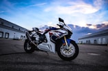 A Honda motorcycle is parked on an expansive, empty asphalt surface, flanked by large industrial buildings under a partly cloudy sky. The bike's design features bold red, white, and blue colors with sleek, aerodynamic lines.