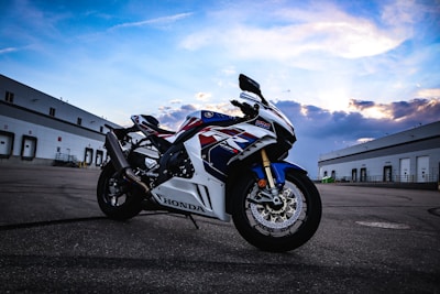 A Honda motorcycle is parked on an expansive, empty asphalt surface, flanked by large industrial buildings under a partly cloudy sky. The bike's design features bold red, white, and blue colors with sleek, aerodynamic lines.
