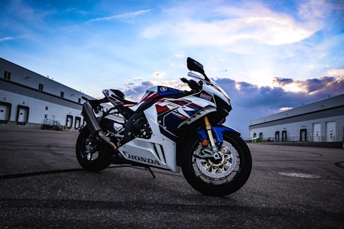 A Honda motorcycle is parked on an expansive, empty asphalt surface, flanked by large industrial buildings under a partly cloudy sky. The bike's design features bold red, white, and blue colors with sleek, aerodynamic lines.