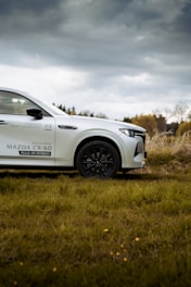 A white Mazda CX-60 plug-in hybrid vehicle parked on grassy terrain, with a cloudy sky in the background. The vehicle's front section is prominently displayed, featuring sleek lines and a modern design. The surroundings include some trees in the distance under an overcast sky.