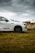 A white Mazda CX-60 plug-in hybrid vehicle parked on grassy terrain, with a cloudy sky in the background. The vehicle's front section is prominently displayed, featuring sleek lines and a modern design. The surroundings include some trees in the distance under an overcast sky.