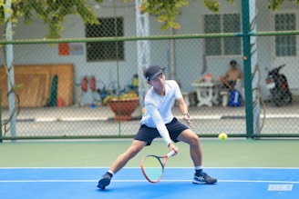 A tennis player dressed in a white shirt and black shorts is making a backhand shot on a blue tennis court. The background shows a chain-link fence with a building, a parked motorcycle, and another person sitting in the shade. The scene is outdoors during daytime, with some greenery visible.