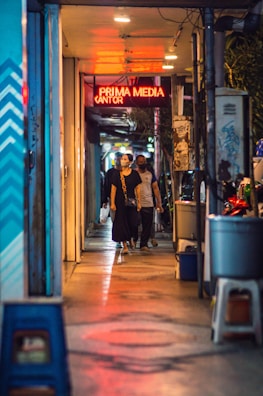 A narrow alleyway with dim lighting features a group of people walking toward the camera. They are under a ceiling with bright, warm-toned lights, forming a contrast with the cool blue tones on the walls. A red neon sign above reads 'PRIMA MEDIA KANTOR'. Various objects like bins and stools are scattered along the sides, and the ground reflects the colorful lights, adding a vibrant touch to the scene.