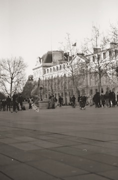 A monochrome photograph capturing a skateboarder in mid-air on a city plaza, with a historical building and leafless trees in the background. A group of people are gathered, some also skateboarding, creating a lively scene.