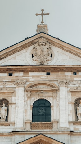 The architectural detail of a historical building facade features intricate stone carvings, a large central arched window with ornate designs, and statues positioned in alcoves. A cross is perched atop the structure, crowning the pediment which is adorned with elaborate decorative elements.