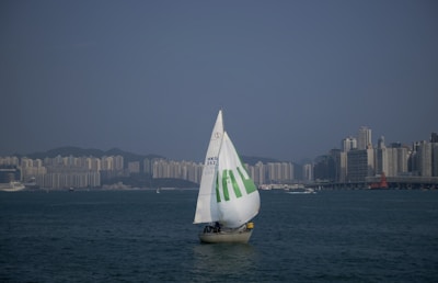 A sailboat with a large white sail marked with green text glides across a broad body of water. The skyline of a city with numerous high-rise buildings is visible on the horizon under a clear blue sky.