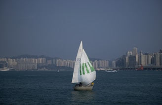 A sailboat with a large white sail marked with green text glides across a broad body of water. The skyline of a city with numerous high-rise buildings is visible on the horizon under a clear blue sky.