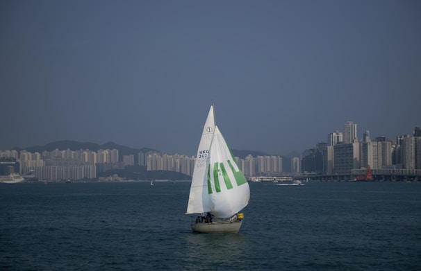 A sailboat with a large white sail marked with green text glides across a broad body of water. The skyline of a city with numerous high-rise buildings is visible on the horizon under a clear blue sky.