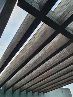 Large wooden beams form an open ceiling, creating a pattern against the backdrop of the sky. The texture of the wood is visible, and the beams are supported by concrete or brick walls.