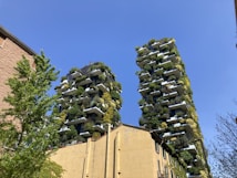 Two modern high-rise buildings covered with abundant greenery, featuring plants on every balcony. The structures are surrounded by a clear blue sky, and other buildings and trees are visible in the foreground.