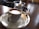 A cup of coffee with a smooth layer of crema sits on a white saucer placed on a silver tray. In the background, a glass cup with a small spoon rests by its side, situated on a wooden table with a blurred, cozy cafe environment.