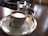 A cup of coffee with a smooth layer of crema sits on a white saucer placed on a silver tray. In the background, a glass cup with a small spoon rests by its side, situated on a wooden table with a blurred, cozy cafe environment.