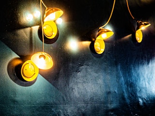 A series of yellow, dome-shaped wall lamps emit warm light against a dark, textured wall. The lamps create circular patterns of illumination with visible points of intense brightness. Wires connect each lamp to the wall, adding a sense of industrial design.
