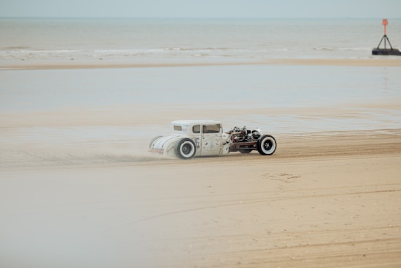 A vintage hot rod car is driving along a sandy beach with the ocean visible in the background. The car appears to be modified with exposed engine parts and large white-walled tires. The setting is overcast, casting a muted light across the scene.