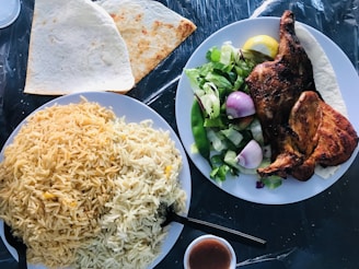 A well-presented meal with a plate of golden-yellow rice, a serving of grilled chicken, and a fresh salad with onions, lettuce, and lemon wedges. Two pieces of flatbread are placed on the side, with a small bowl of sauce nearby.