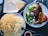 A well-presented meal with a plate of golden-yellow rice, a serving of grilled chicken, and a fresh salad with onions, lettuce, and lemon wedges. Two pieces of flatbread are placed on the side, with a small bowl of sauce nearby.