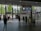 A train station interior with people walking and interacting near ticket gates. Information boards display time and departure details. Natural light filters through large glass windows, providing a view of trees and modern buildings outside.