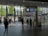 A train station interior with people walking and interacting near ticket gates. Information boards display time and departure details. Natural light filters through large glass windows, providing a view of trees and modern buildings outside.