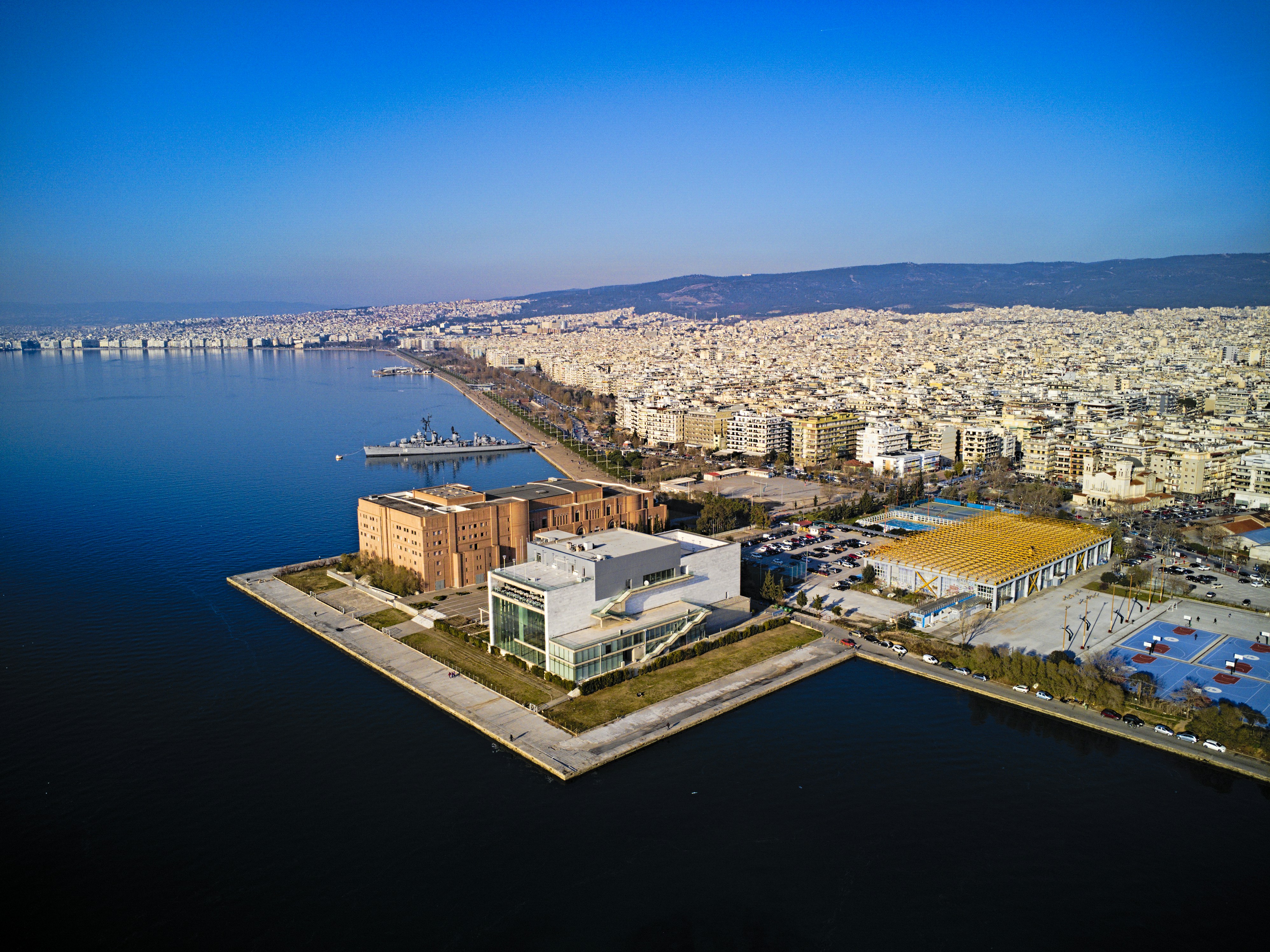 a large body of water with a city in the background, This aerial drone photo captures the stunning waterfront of Thessaloniki, Greece, with the iconic concert hall and historic warship "Velos" visible in the foreground. The crystal-clear waters of the Aegean Sea lap at the city