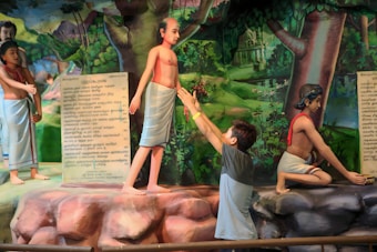 A scene featuring a series of statues depicting historical or religious figures in traditional attire. The background is painted with a lush forest landscape. One male figure stands with hands outstretched towards the statues, appearing to be in a gesture of offering or reverence. Another statue is kneeling, while a third stands near a stone tablet inscribed with text.
