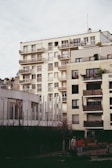 Several modern multi-story apartment buildings with balconies, featuring a blend of white, beige, and gray tones. The foreground includes a small garden area with a couple of indistinct figures and some outdoor seating.