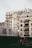 Several modern multi-story apartment buildings with balconies, featuring a blend of white, beige, and gray tones. The foreground includes a small garden area with a couple of indistinct figures and some outdoor seating.