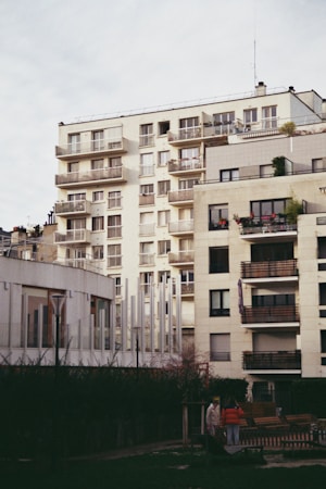 Several modern multi-story apartment buildings with balconies, featuring a blend of white, beige, and gray tones. The foreground includes a small garden area with a couple of indistinct figures and some outdoor seating.