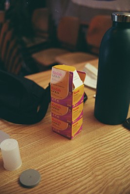 A wooden table holds several boxes of Kodak Gold film stacked vertically. Nearby are a black bottle and a black bag, along with a couple of plastic film canisters and their lids.
