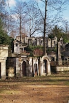 A cemetery scene with ornate stone tombs and mausoleums surrounded by tall leafless trees. The structures show signs of aging, with some walls covered in vines. There is a grassy and dirt foreground with scattered leaves, creating a serene but slightly somber atmosphere.