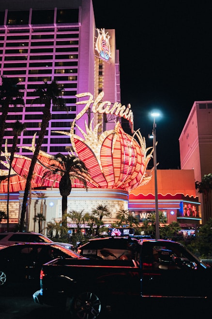 A brightly lit casino with neon lights in vibrant red, pink, and blue, featuring a large flamingo signage. Palm trees are visible in the foreground with vehicles passing by on the street at night.