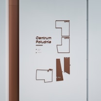 A wall-mounted directory map with text and abstract geometric shapes in brown against a light blue background. The words 'Centrum Południe' are prominently displayed alongside location indicators labeled 'CP1', 'CP2', 'CP3', and 'CP4'. Small text below the main title includes 'jesteś tutaj' and 'wejście'.